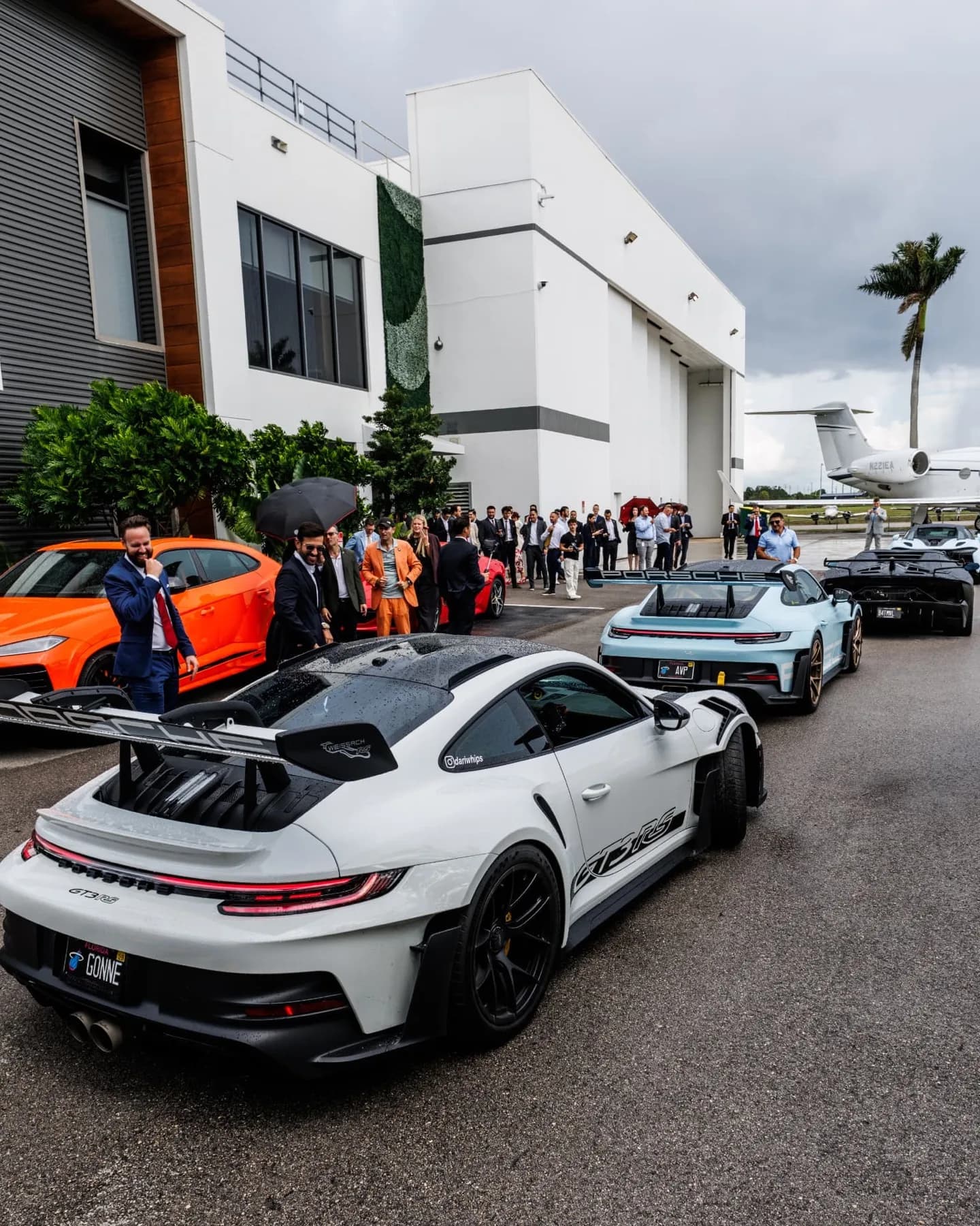 High angle view of supercars parked outside a white hangar building with a private jet behind