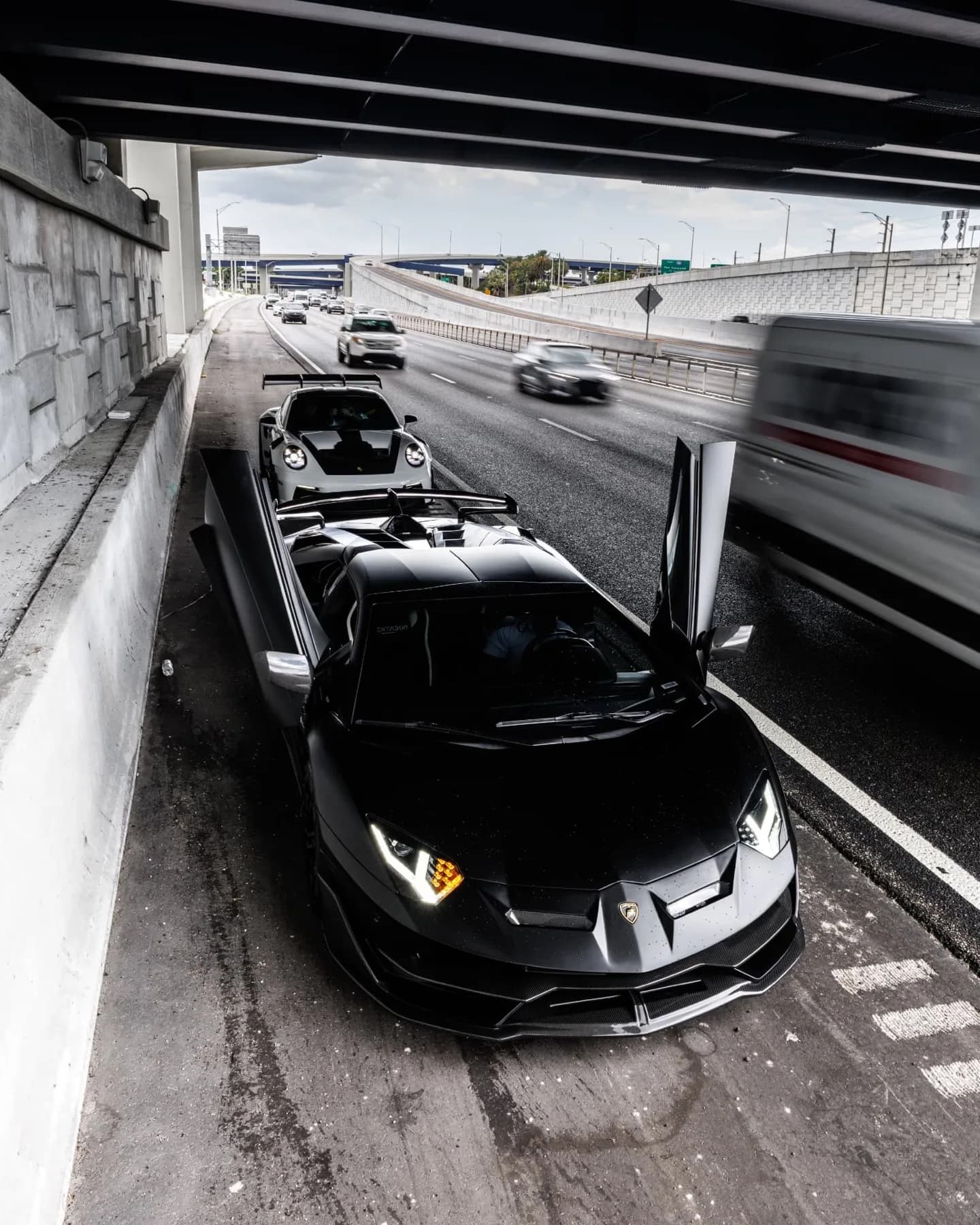 Black Lamborghini Aventador SVJ and white Porsche GT3 RS parked together under a highway overpass