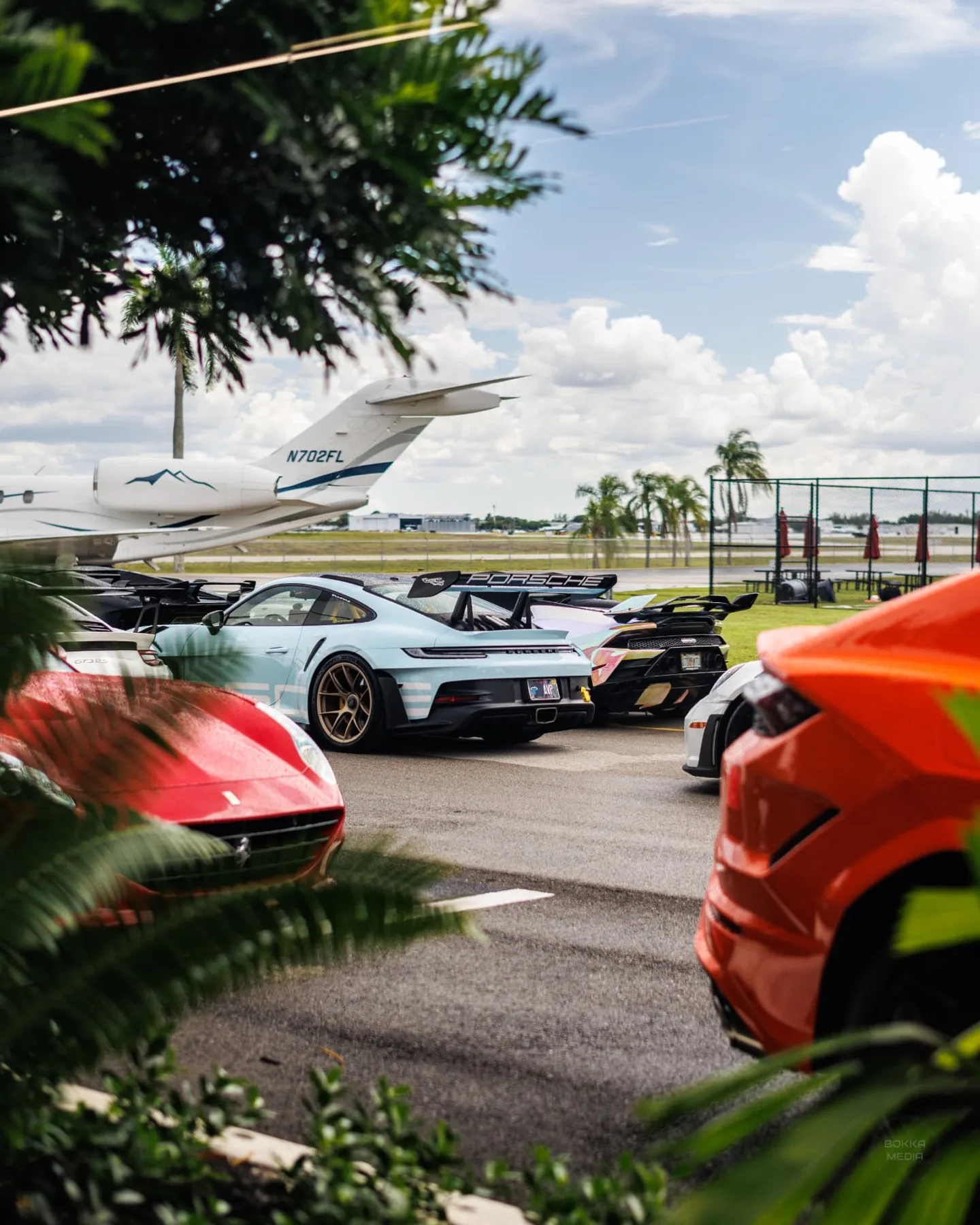 Angelo's Miami blue Porsche GT3 RS and an iridescent Lamborghini seen through tropical foliage with a jet behind