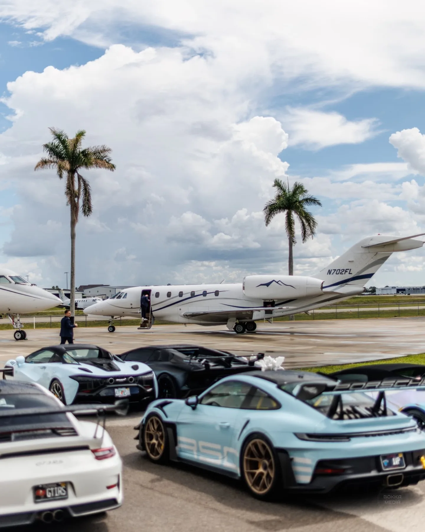 Wide view of supercars and private jets on the ramp under a cloudy Florida sky