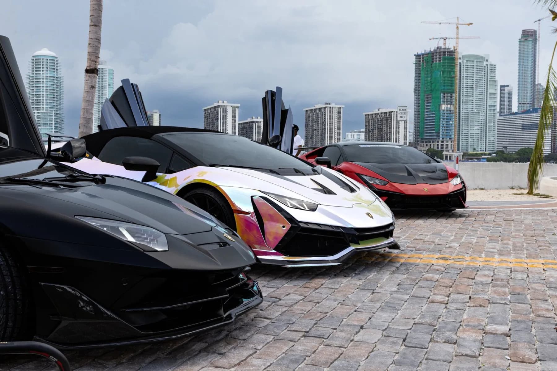 Lamborghinis and a Ferrari parked with doors raised along the waterfront at the end of the drive.